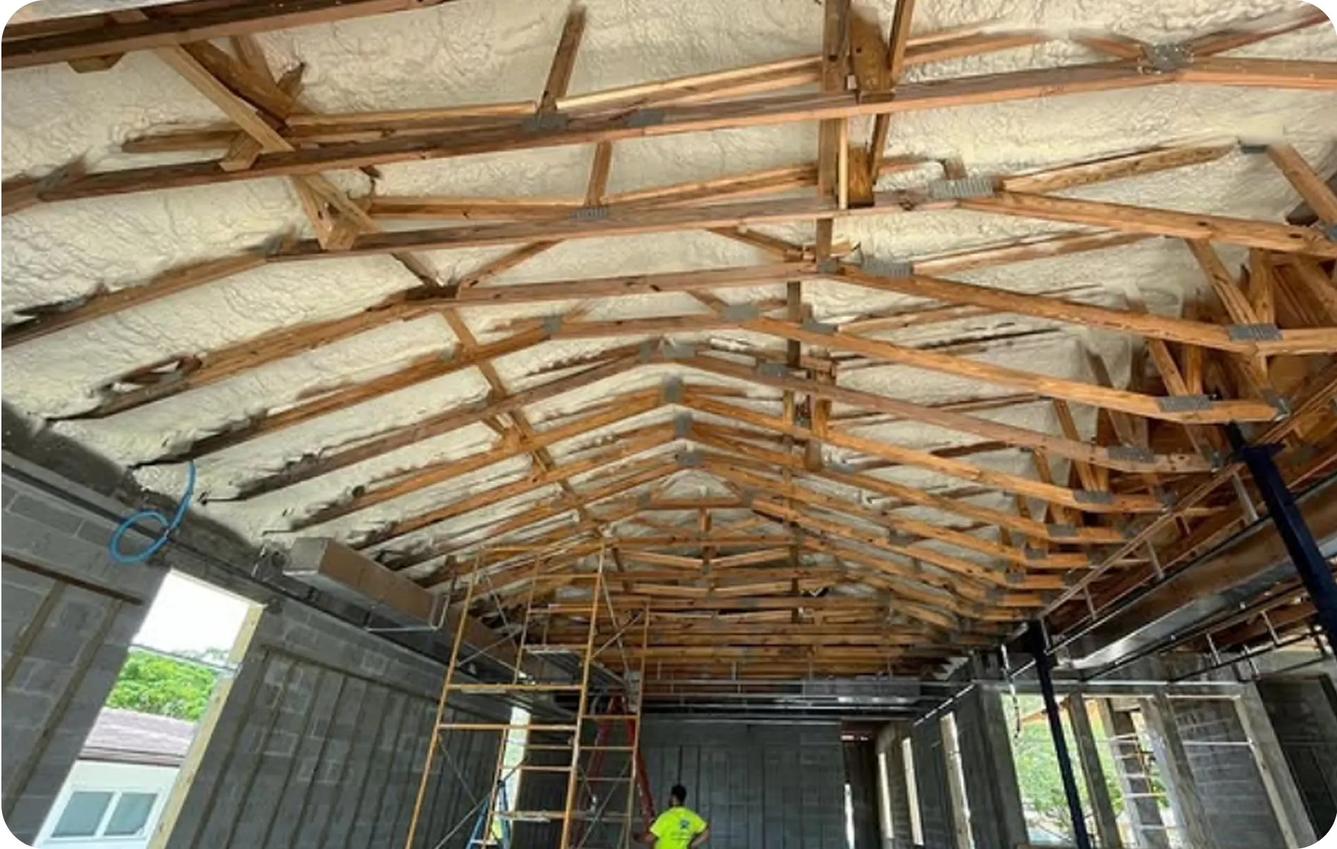 Spray foam insulation applied to the underside of a roof deck between timber trusses, over block walls in a building under construction