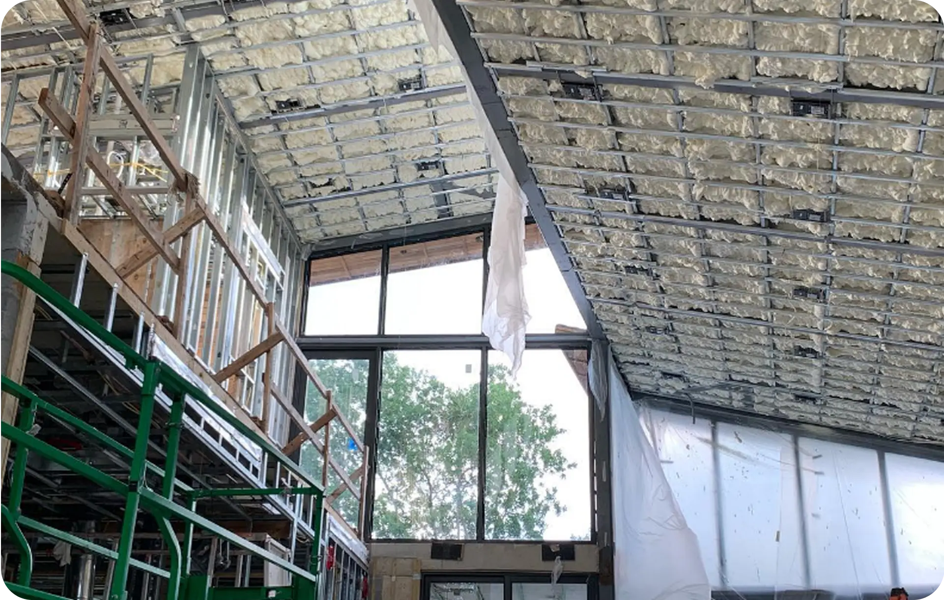 Spray foam insulation filling the bays between steel framing on a sloped ceiling, beside a full-height window wall on a commercial job